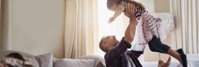 Australia’s most trusted health fund. Man wearing glasses sitting on white couch lifting young girl in the air in playful manner