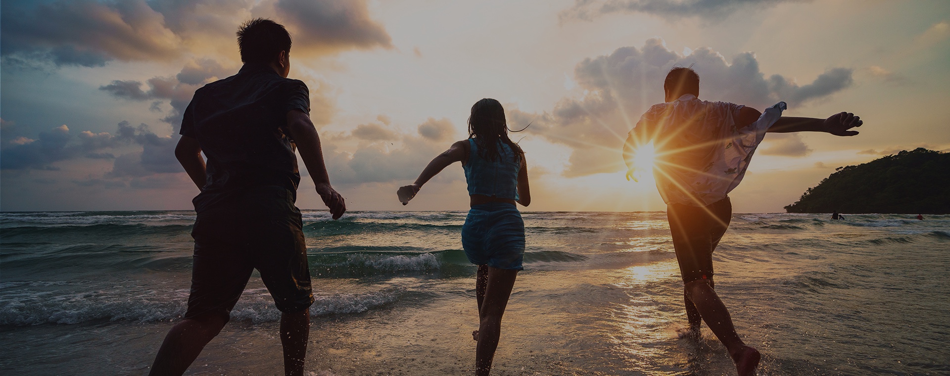 Two teenage boys and young girl running into the shallow ocean with golden sun