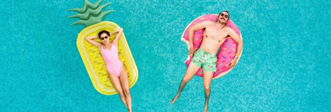 Treat yourself more often Young woman in pink swimsuit on a yellow pineapple shaped lilo next to man with green trucks on a pink donut shaped lilo in a pool