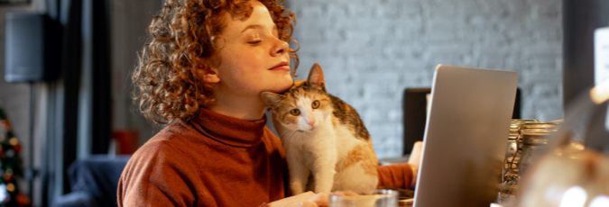 Woman with curly hair rests chin on cat whilst gazing at laptop screen
