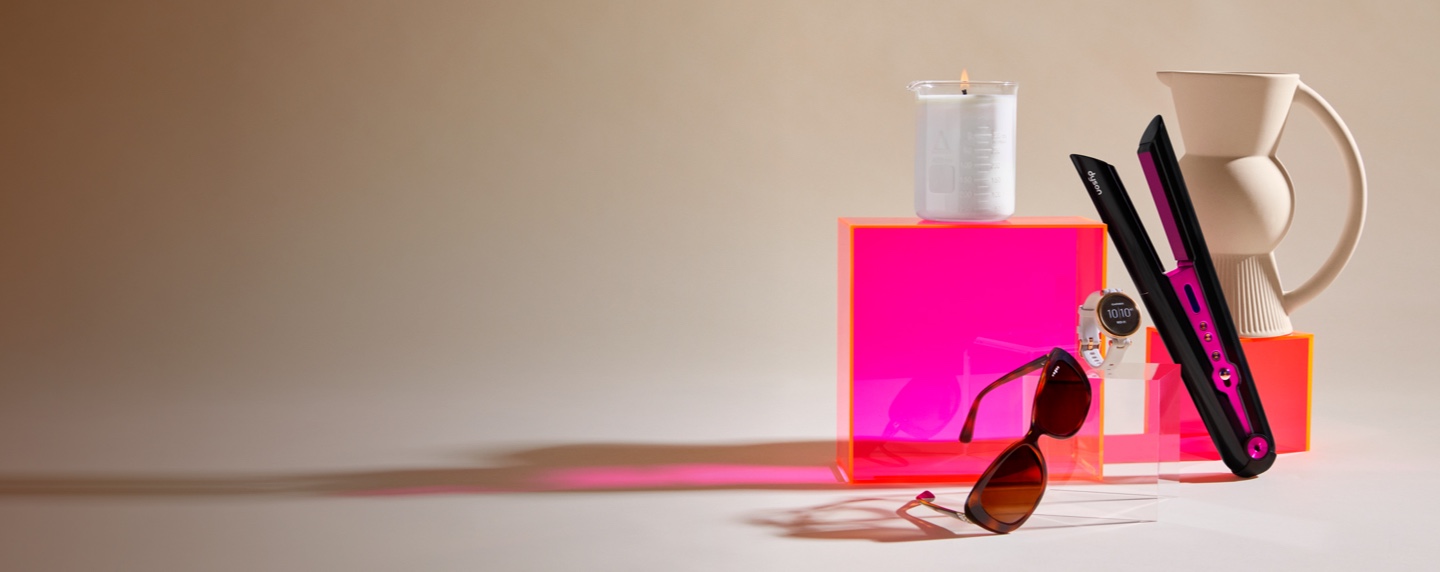 Eggshell coloured backdrop with fluoro pink boxes displaying a clear beaker candle, vogue brown sunglasses, Garmin ladies sportswatch, pink and black hair straightener and eggshell coloured jug with fan detail.