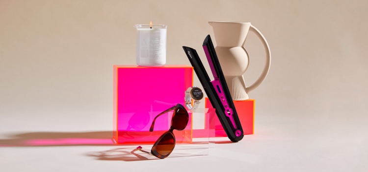 Eggshell coloured backdrop with fluoro pink boxes displaying a clear beaker candle, vogue brown sunglasses, Garmin ladies sportswatch, pink and black hair straightener and eggshell coloured jug with fan detail.