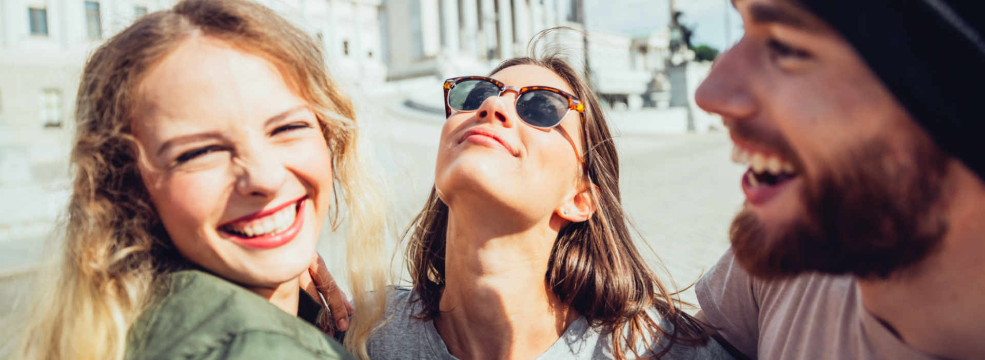 Smiling blonde women next to women with sunglasses looking at the sky and bearded man with beanie outside grand building